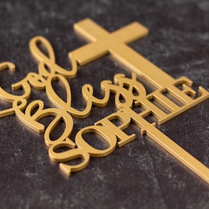 Pu&ograve; includere: Un topper per torta color oro caratterizzato da una forma a croce integrata con il testo God Bless SOPHIE in una combinazione di caratteri corsivi e stampatello. Il topper include un lungo e sottile picchetto verticale per l'inserimento nel dolce.