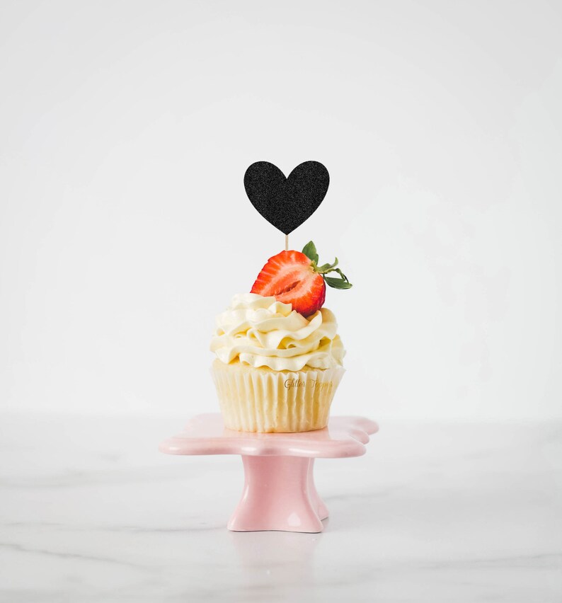 Op de afbeelding: Een enkele cupcake met witte frosting en een aardbei erop. Een zwarte hartvormige topper zit in de aardbei. De cupcake staat op een roze taartstandaard.