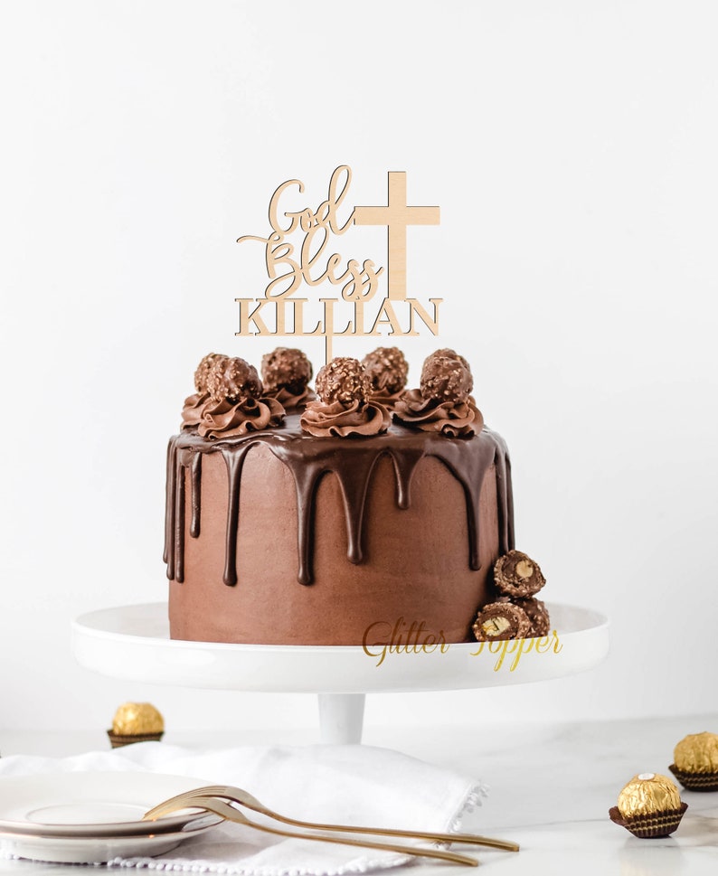 Pu&ograve; includere: Un topper per torta in legno chiaro con una croce e la scritta God Bless KILLIAN, posizionato sopra una torta al cioccolato rotonda con glassa al cioccolato fondente, guarnita con ciuffi di crema al cioccolato e cioccolatini alla nocciola.