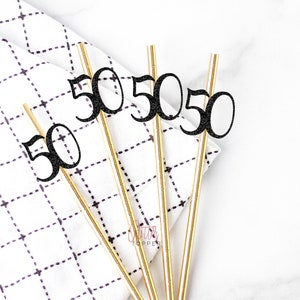 May include: Four gold straws with black glitter 50s on top. The straws are on a white and black checkered fabric.