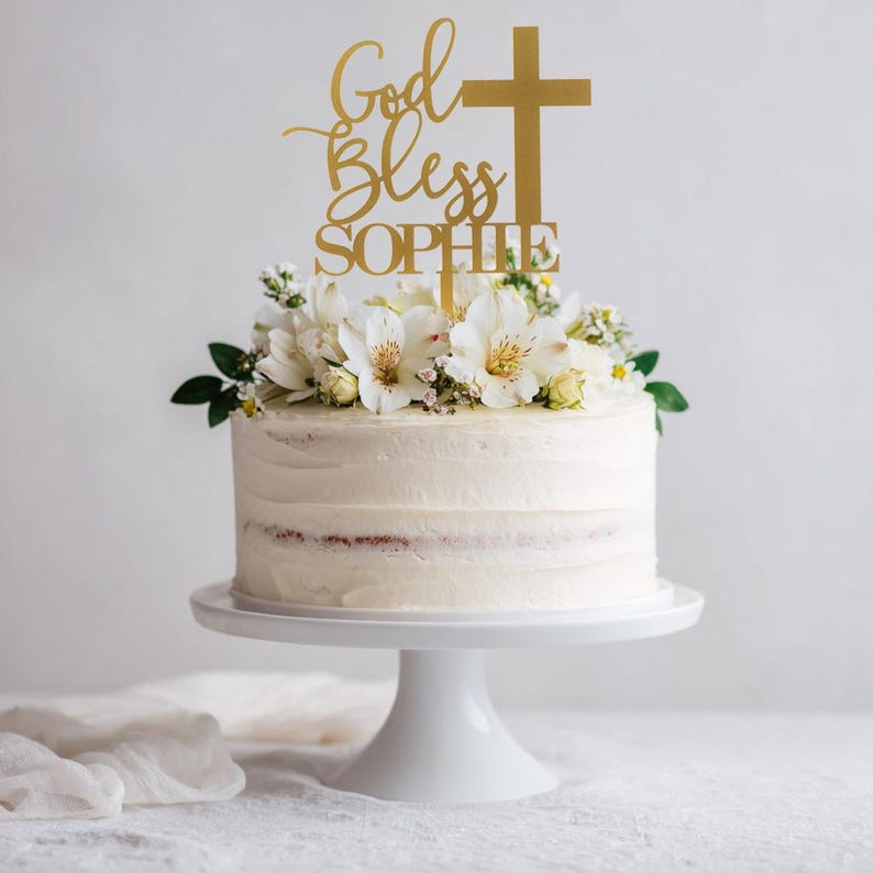 Pu&ograve; includere: Un topper per torta color oro con la scritta God Bless SOPHIE e il simbolo di una croce, posizionato sopra una torta con glassa bianca decorata con fiori bianchi. Il topper &egrave; realizzato in un materiale sottile e piatto, inserito al centro.