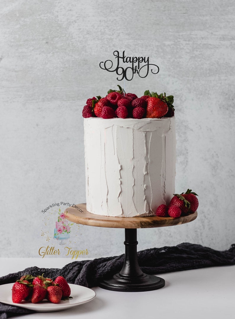 May include: White cake with a black glitter topper that says "Happy 90th" and topped with fresh strawberries and raspberries. The cake is on a wooden cake stand with a black base.