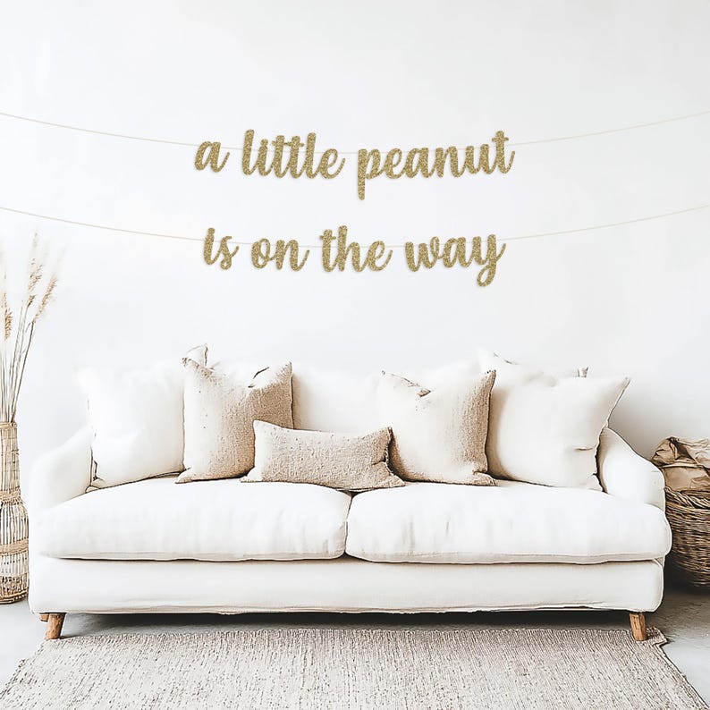 Puede incluir: Una pancarta de brillo dorado con el texto "a little peanut is on the way" colgada en una pared blanca sobre un sof&aacute; blanco con almohadas.