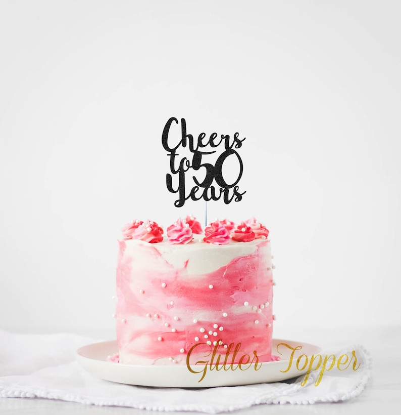 May include: Black glitter cake topper with the text "Cheers to 50 Years" on a pink and white frosted cake.