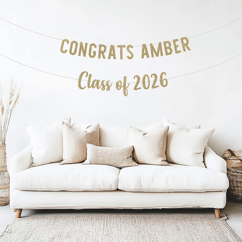 May include: A gold glitter banner that reads "CONGRATS AMBER" and "Class of 2026" hangs above a white couch with beige pillows. The banner is strung on a thin white string, perfect for a graduation celebration.