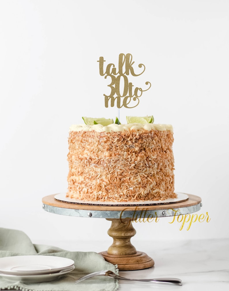 May include: A white cake decorated with shredded coconut and lime slices, topped with a gold glitter cake topper that reads "talk 30 to me".
