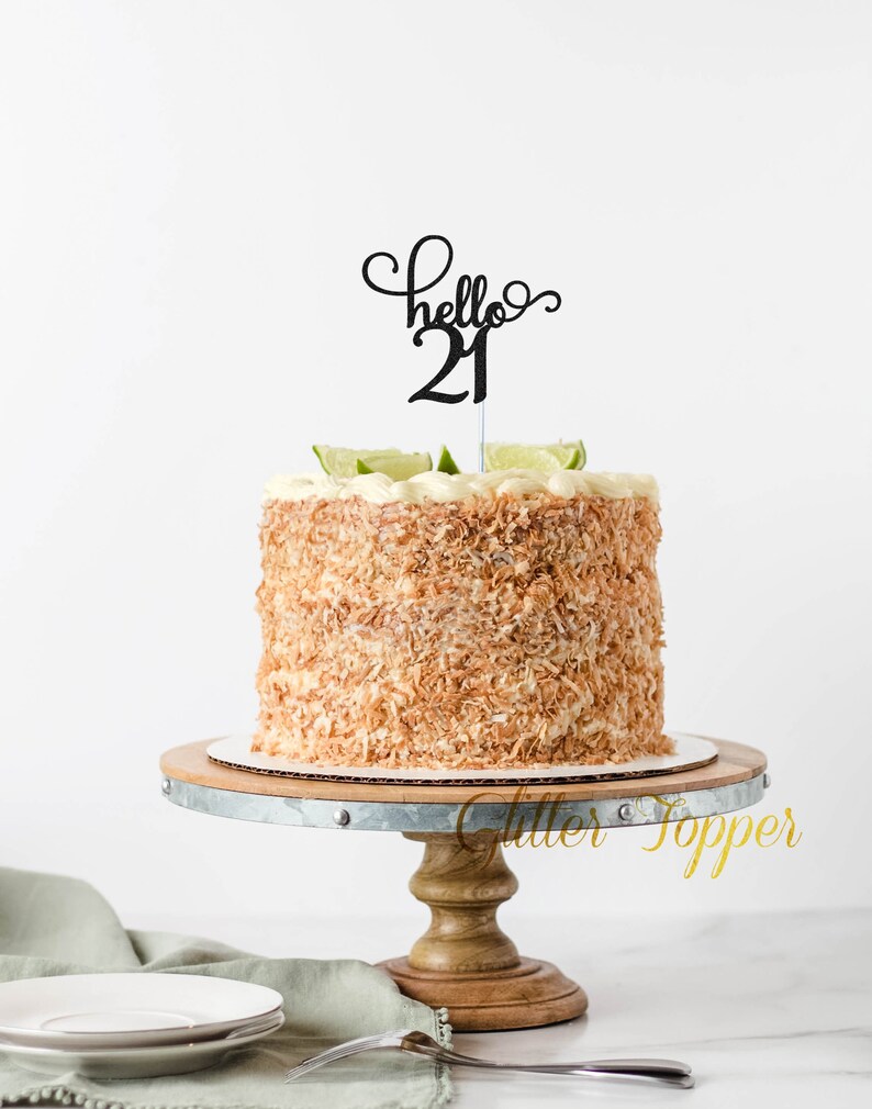 May include: A white cake with coconut flakes and lime slices on top, decorated with a black cake topper that says "hello 21". The cake is on a wooden cake stand with a metal rim.