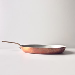 May include: A copper-colored frying pan with a long, slender handle. The pan has a rounded shape with a silver interior. The handle is a darker brown color. The pan is sitting on a white surface.