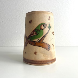 May include: A beige ceramic mug with a brown and green painted bird on a branch. The mug has a brown rim and base.