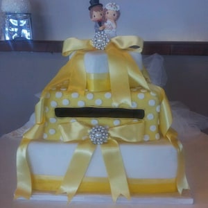 Op de afbeelding: Een drielaagse bruidstaart met geel lint en witte stippen. De bovenste laag heeft een klein bruidspaarfiguurtje erop.