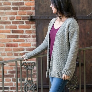 May include: A gray crocheted cardigan with a V-neck and long sleeves. The cardigan is open in the front and is being worn over a maroon shirt.