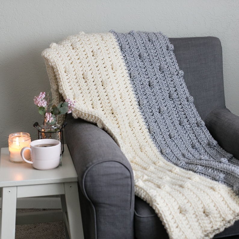 May include: A white and grey crocheted throw blanket with a textured bobble stitch pattern draped over a grey armchair.