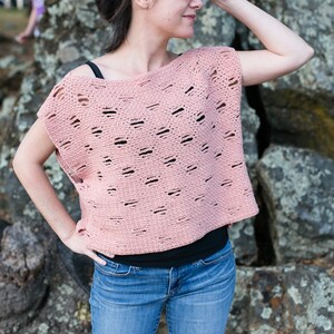 Puede incluir: Un top de crochet rosa con un diseño suelto y tejido abierto. El top tiene un escote redondo ancho y mangas cortas. La modelo lleva el top con jeans azules.