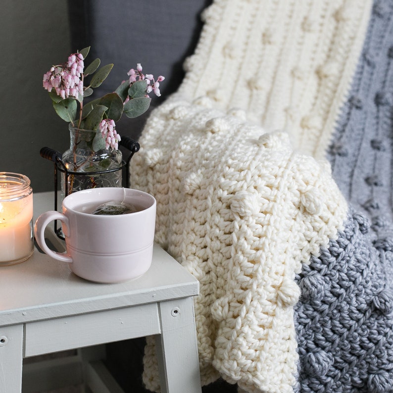 May include: A white crocheted throw blanket with a textured pattern and a grey crocheted border. The blanket is draped over a chair and a cup of tea is on a white table in the foreground.