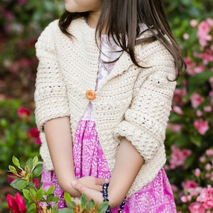 May include: A young girl wearing a cream crocheted cardigan with a pink floral dress and a beaded necklace. The cardigan has a button closure and the girl is standing in front of a pink flower bush.