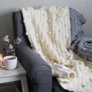 May include: A white and grey crocheted throw blanket with a textured bobble stitch pattern draped over a grey armchair.