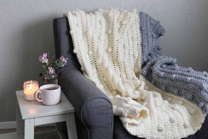 May include: A white and grey crocheted throw blanket with a textured bobble stitch pattern draped over a grey armchair.