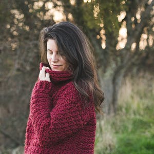 May include: A woman wearing a deep red crocheted sweater with a turtleneck collar. The sweater has a textured stitch pattern and is loose fitting.
