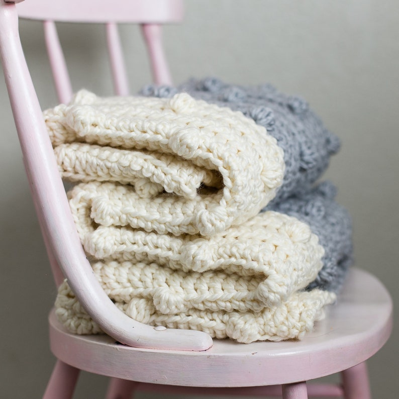 May include: Two crocheted blankets, one white and one grey, are folded and stacked on a pink wooden chair. The white blanket has a textured, bobbly surface.