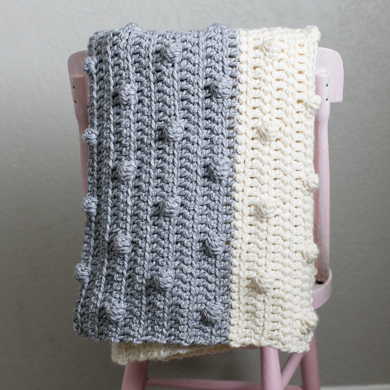 May include: A grey and white crocheted baby blanket with a textured bobble stitch. The blanket is draped over a pink chair.