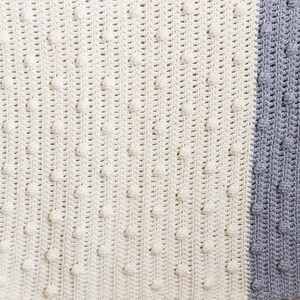 May include: A crocheted baby blanket in a white and grey colour scheme. The blanket has a textured pattern with raised bumps.