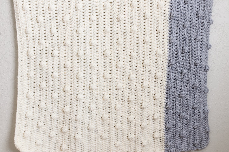 May include: A crocheted baby blanket in a white and grey colour scheme. The blanket has a textured pattern with raised bumps.