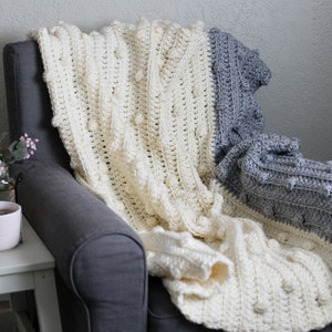 May include: A white and grey crocheted throw blanket with a textured bobble stitch pattern. The blanket is draped over a grey armchair.