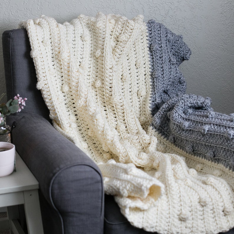 May include: A white and grey crocheted throw blanket with a textured bobble stitch pattern. The blanket is draped over a grey armchair.