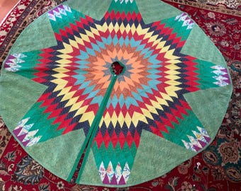 May include: A large, round Christmas tree skirt with a starburst design. The skirt features a green background with a central star pattern in shades of red, orange, yellow, blue, and green. The skirt has a slit for the tree trunk.