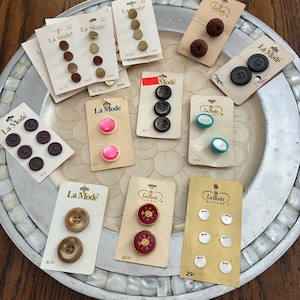 May include: Assortment of vintage "La Mode" buttons on their original cards. The buttons vary in color and style, including brown, black, pink, and iridescent options. The cards display prices and style numbers.