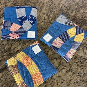 May include: Three blue quilted coasters with various patterned fabric accents. Each coaster features a different design, including nautical anchors, stripes, and geometric patterns. The coasters are numbered 1, 2, and 3 on small white labels.