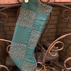 May include: A handmade Christmas stocking with a patchwork design. The stocking features squares of solid green fabric and fabric with a floral pattern in shades of yellow, orange, and white on a dark background. A white button and loop are at the top.