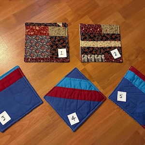 May include: Five square fabric pot holders arranged in a group. Two feature patchwork patterns in red, black, and beige. Three feature solid blue, red, and light blue stripes with quilted stitching. Each item has a small white paper label numbered 1 to 5.