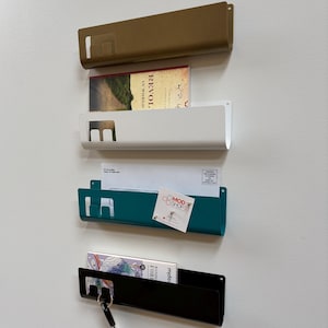 May include: Four wall-mounted metal organizers in gold, white, teal, and black. Each features a rectangular slot on the left side for hanging items like keys. The organizers hold various items including a book, envelopes, and a small business card.