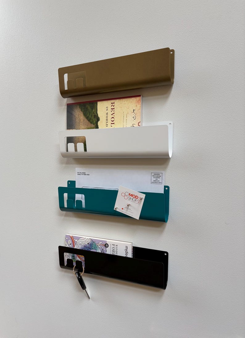 May include: Four wall-mounted metal organizers in gold, white, teal, and black. Each features a rectangular slot on the left side for hanging items like keys. The organizers hold various items including a book, envelopes, and a small business card.