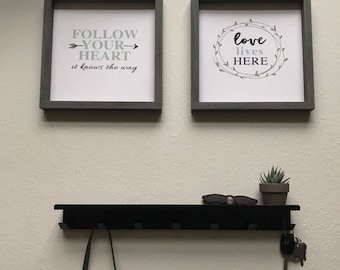 May include: Two framed prints with inspirational quotes hang on a white wall above a black shelf with hooks. The left print says "Follow your heart it knows the way". The right print says "Love lives here" with a wreath of leaves around the words. A small potted succulent plant sits on the shelf.