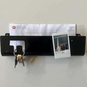 May include: A black wall-mounted mail organizer with a horizontal slot holding a white envelope from Target Card Services. The organizer features two integrated hooks on the left side holding a metal keyring and a small square photograph on the right.