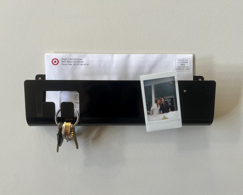 May include: A black wall-mounted mail organizer with a horizontal slot holding a white envelope from Target Card Services. The organizer features two integrated hooks on the left side holding a metal keyring and a small square photograph on the right.