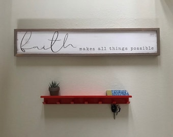 May include: A rectangular wooden sign with the word "Faith" in cursive and the phrase "makes all things possible" in a serif font. Below, a red shelf with pegs holds a small potted plant, keys, and a decorative item.