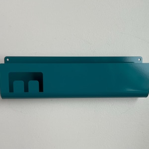 May include: A teal metal wall-mounted organizer featuring a rectangular shape with a smooth finish. The front panel includes a cutout design resembling a lowercase letter m on the left side, and two small circular holes for mounting at the top.