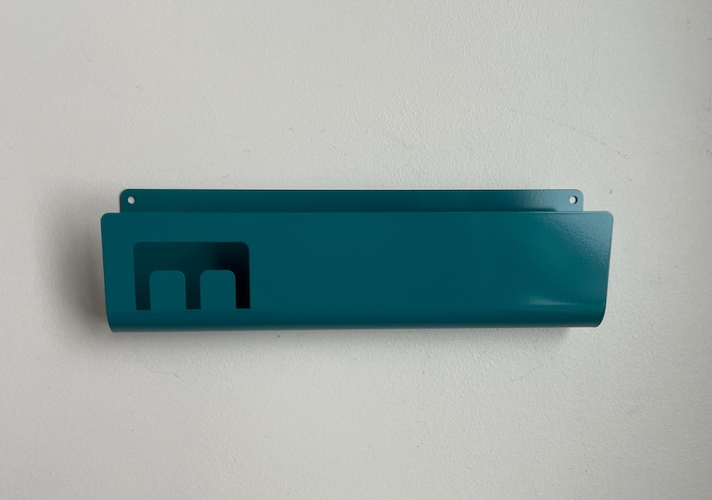 May include: A teal metal wall-mounted organizer featuring a rectangular shape with a smooth finish. The front panel includes a cutout design resembling a lowercase letter m on the left side, and two small circular holes for mounting at the top.
