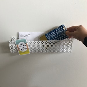 May include: A white, geometric-patterned wall-mounted basket holding a card that says "It's Gonna Be OK", a white envelope, and a blue and gold phone case. The basket is mounted on a white wall.