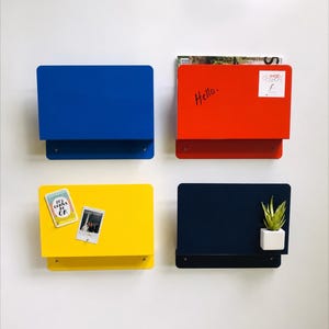 May include: Four rectangular wall-mounted metal organizers in blue, orange, yellow, and dark navy. The orange unit features the handwritten text Hello and a Mod Shop sticker. The yellow unit holds two small cards, and the navy unit holds a small plant.
