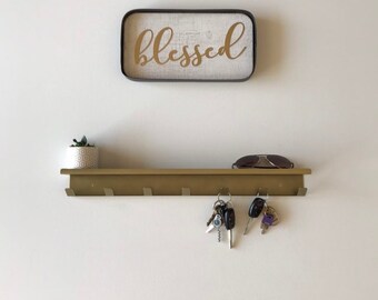 May include: A gold metal key rack with five hooks, mounted on a white wall. The rack has a shelf above it. A small potted plant sits on the shelf, and a pair of sunglasses are resting on the shelf. A set of keys hangs from one of the hooks. A sign with the word "blessed" in gold lettering is hanging on the wall above the shelf.