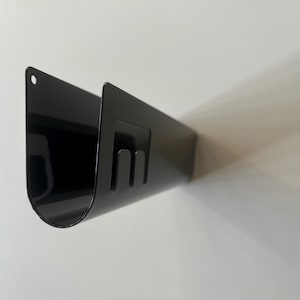 May include: A black U-shaped metal wall mount organizer with a smooth, glossy finish. The piece features a rounded bottom, a small circular hole at the top left corner for mounting, and a rectangular cutout section on the side with two prongs.
