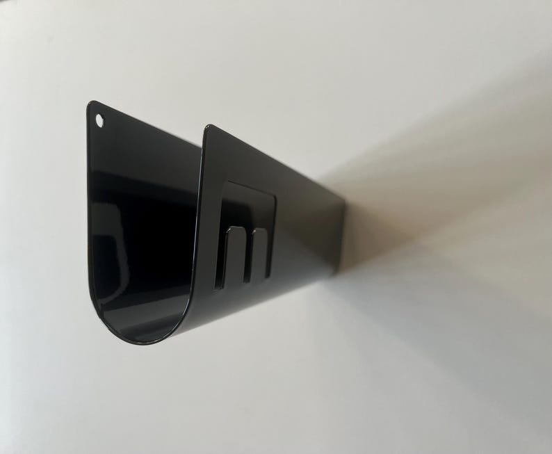 May include: A black U-shaped metal wall mount organizer with a smooth, glossy finish. The piece features a rounded bottom, a small circular hole at the top left corner for mounting, and a rectangular cutout section on the side with two prongs.