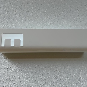 May include: A rectangular white wall-mounted holder with a smooth, glossy finish. The object features a cutout section on the left side that reveals a distinct letter E shape, providing an open space for storage or display purposes.