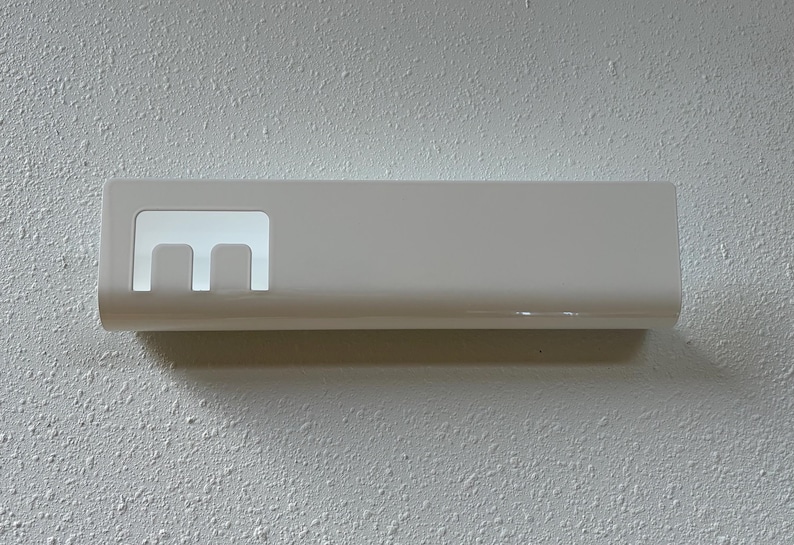 May include: A rectangular white wall-mounted holder with a smooth, glossy finish. The object features a cutout section on the left side that reveals a distinct letter E shape, providing an open space for storage or display purposes.