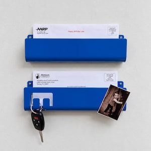 May include: Two blue metal wall-mounted mail organizers with a smooth finish. The top holder contains an AARP envelope. The bottom holder features a cutout for hanging keys, holds a Shriners Children's envelope, and displays a small photograph.