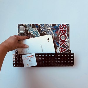 May include: A black metal wall-mounted rack with a perforated grid pattern. The rack holds a magazine titled SPOKA with a blue wave graphic and a white Samsung Galaxy Tab tablet. A small square card with the text MOD SHOP GIRL is attached.